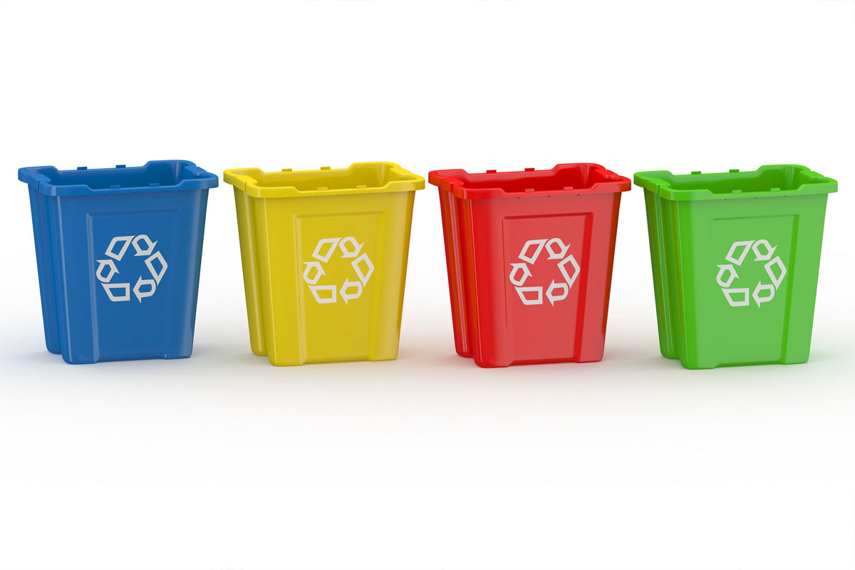 Immagine con quattro contenitori per la raccolta differenziata allineati su sfondo chiaro. I bidoni sono di colore blu, giallo, rosso e verde e riportano sul lato frontale il simbolo bianco del riciclo.
