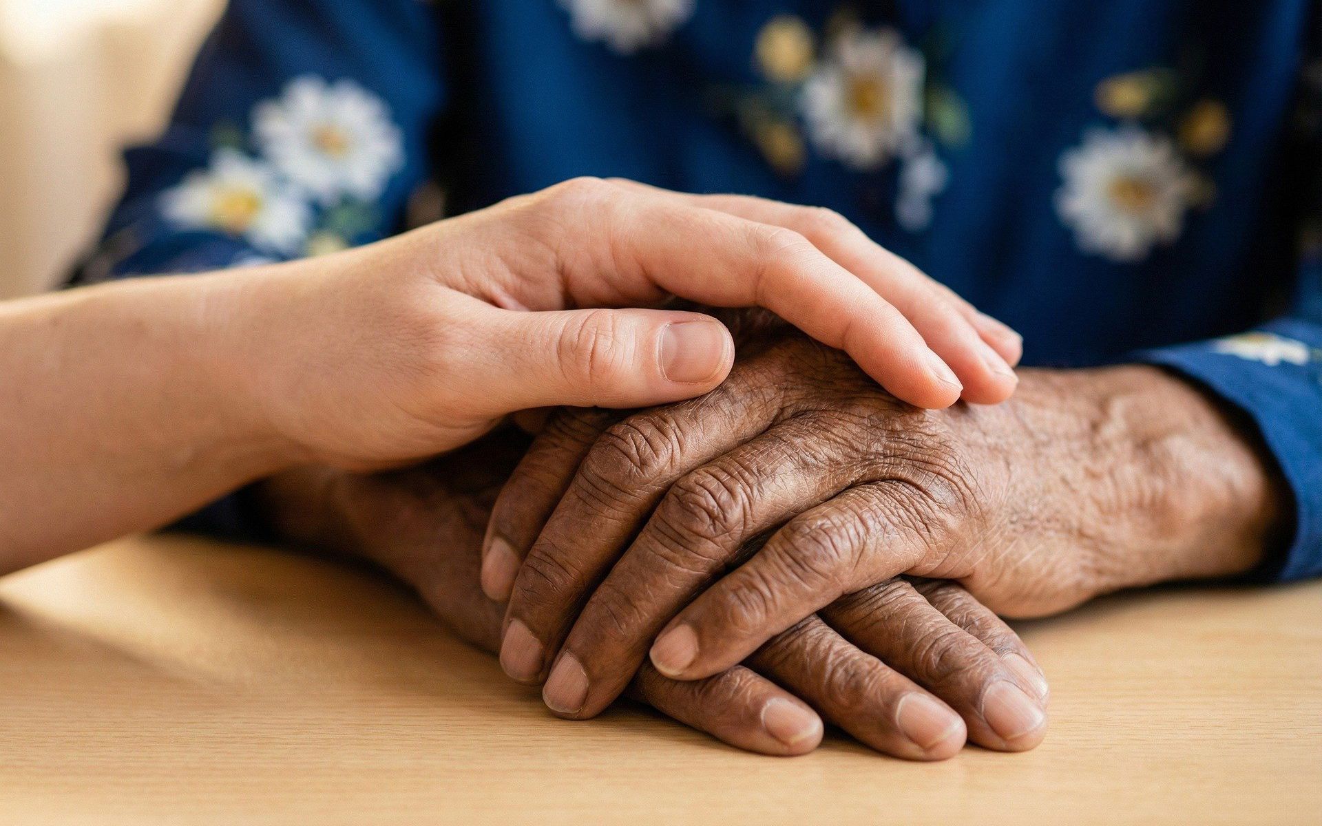 In primo piano, su un tavolo di legno, sono raffigurate le mani di due persone. Una mano più giovane e dalla pelle chiara appoggia delicatamente il palmo sul dorso della mano di una persona anziana, dalla pelle scura e segnata dalle rughe. Le mani dell’anziano sono raccolte e sovrapposte sul tavolo. Sullo sfondo, sfocata, si intravede una persona che indossa un abito blu con motivi floreali.